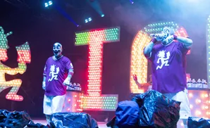 Performers in clown makeup and purple jerseys on a stage with lit signs and colorful lights.