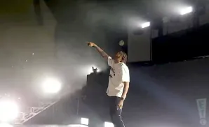 A man in a white t-shirt and black pants stands on a stage, pointing upwards into smoke and stage lights.