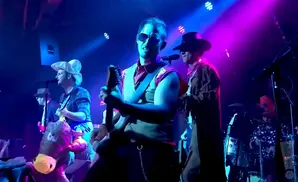Band members in cowboy hats perform on stage, one playing guitar and another at the drums.
