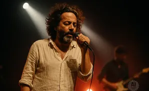 A man with curly hair and a beard sings into a microphone on a dimly lit stage, with a guitarist blurred in the background.