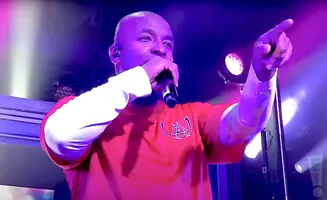 A bald Black man in an orange shirt points forward while holding a microphone.
