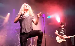 A male singer with long blonde hair performs on stage with a guitarist in the background.