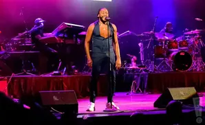 A singer in a dark vest and pants performs on stage with a band.