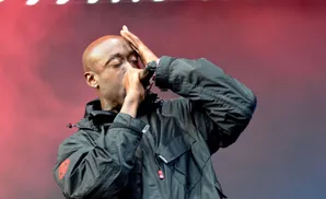 Freddie Gibbs performs on stage, wearing a black jacket and holding a microphone to his mouth with his hand on his forehead.