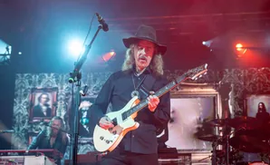 A male guitarist in a black hat and shirt plays a white electric guitar on stage with red and blue lighting.