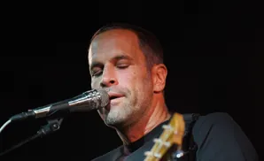A man plays an acoustic guitar and sings into a microphone on a stage with blue lighting.