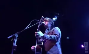 A man on stage plays an acoustic guitar and sings into a microphone, bathed in blue and purple light.