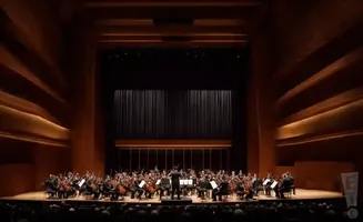An orchestra plays on a stage with wooden paneling and a dark curtain backdrop.