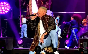 A male singer on stage wearing a long patterned jacket and blue pants, holding a microphone.