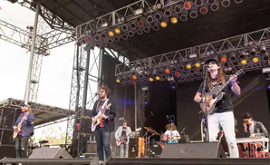 A band plays on an outdoor stage with a large lighting rig overhead and many speakers on the ground.