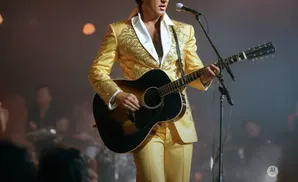 A performer in a gold jumpsuit plays a guitar on stage, with a microphone in front of him.