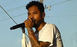 Singer Miguel performing on stage, holding a microphone and wearing a white t-shirt.