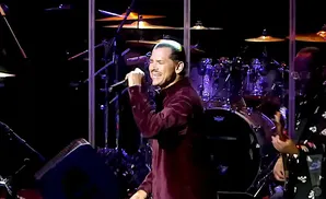 A man in a maroon shirt sings into a microphone on a stage with drums behind him.