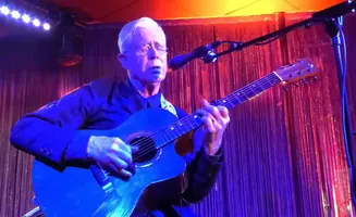 Man with white hair and glasses plays an acoustic guitar on stage under blue and pink lights.