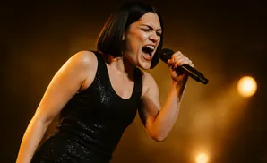 A woman with short dark hair sings into a microphone on a dimly lit stage.