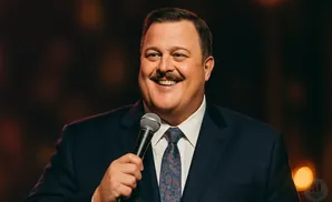 Comedian Billy Gardell smiles with a mustache and holds a microphone.