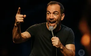 A man with a beard points with his left index finger while holding a microphone and speaking