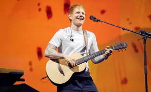 Ed Sheeran plays an acoustic guitar on stage with an orange backdrop.
