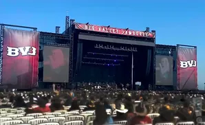 A large outdoor concert stage with screens showing performers, banners, and a crowd in the foreground.