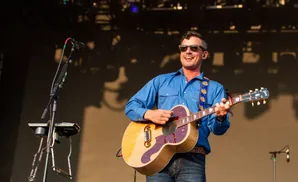 A man in sunglasses and a blue shirt plays an acoustic guitar on stage.