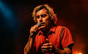 A man with blond, wavy hair sings into a microphone on a dark stage with red and blue lights.