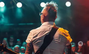 Guitarist with tattoos and spiky hair on stage under stage lights, with audience in the background.