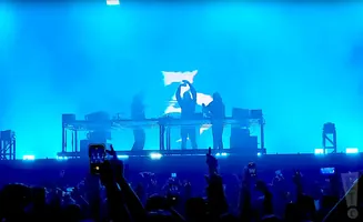 A DJ trio performs on a stage bathed in blue light, with a crowd in the foreground holding up phones.
