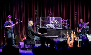 Musicians perform on a stage with a piano, drums, bass, and mandolin.