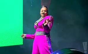 A woman in a bright pink jumpsuit and jacket with plaid trim sings on stage, gesturing with her hand.