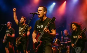 A rock band performs on stage with a male singer in the foreground playing a red guitar.