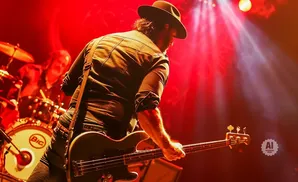A man in a hat plays a bass guitar on a stage with red lighting.