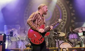 A musician plays a red electric guitar on stage, with a drummer in the background.