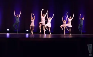 Dancers in pink dresses and one man in a grey shirt and black pants perform on a stage with a purple backdrop.