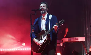 A man with a beard and a blue pinstripe suit sings into a microphone while playing an acoustic guitar on stage.