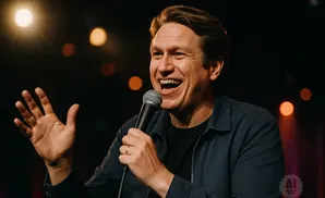A smiling man with brown hair holds a microphone and gestures with his left hand.