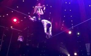 Performers balance on a tightrope with one man on another's shoulders, while a third juggles a bike wheel.