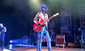 A man in a hat plays a red electric guitar on a dimly lit stage.