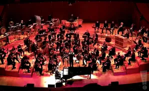 An orchestra plays on a tiered stage under red lighting, with a conductor leading from the front.