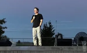 A man in a black t-shirt and khaki pants speaks into a microphone on a stage, with a large fan behind him.