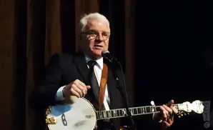 Steve Martin plays a banjo while singing into a microphone.