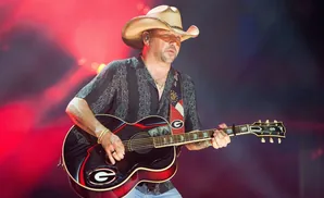 A man in a cowboy hat plays an acoustic guitar on stage with red and blue lights.
