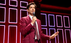 Comedian John Mulaney in a red suit on stage with a microphone, speaking to an audience.