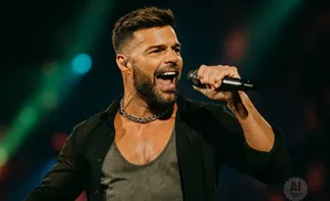 Ricky Martin sings into a microphone on stage, wearing a black open shirt over a gray tank top and a thick chain necklace.