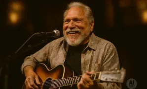 A smiling man with gray hair and a beard plays an acoustic guitar in front of a microphone.