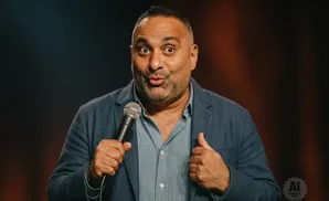 Comedian Russell Peters holding a microphone and giving a thumbs up on stage.