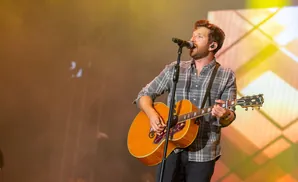 A male singer with reddish-brown hair, wearing a plaid shirt, sings into a microphone while playing an acoustic guitar on stage.