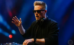 DJ wearing sunglasses and headphones mixes music at a console with blue and red lights.