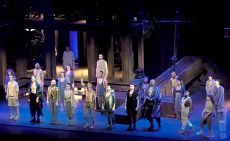 A diverse cast stands on stage, dressed in tunics and flowing robes, under dramatic lighting.