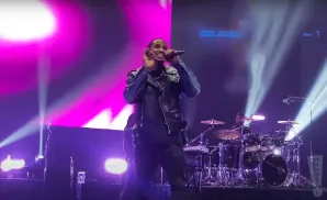 A singer in a leather jacket holds a microphone on stage with a drummer behind him.