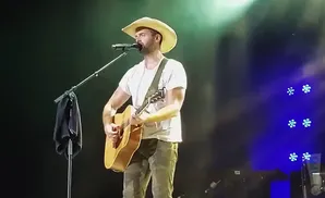 A male singer in a cowboy hat plays an acoustic guitar and sings into a microphone on a dark stage with blue lights.
